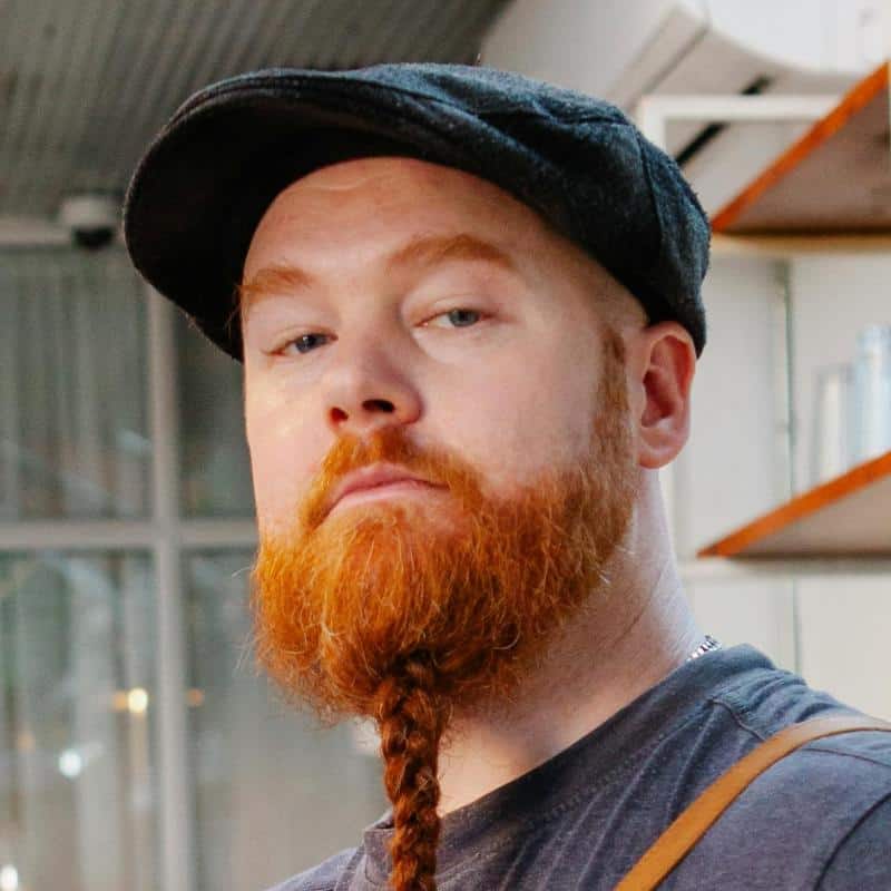 A person with a red beard styled in a braid, wearing a flat cap and a dark shirt, looks at the camera in an indoor setting.