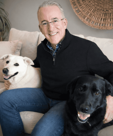 A man wearing glasses and a black sweater sits on a couch, smiling, with a white dog on his left and a black dog on his right.