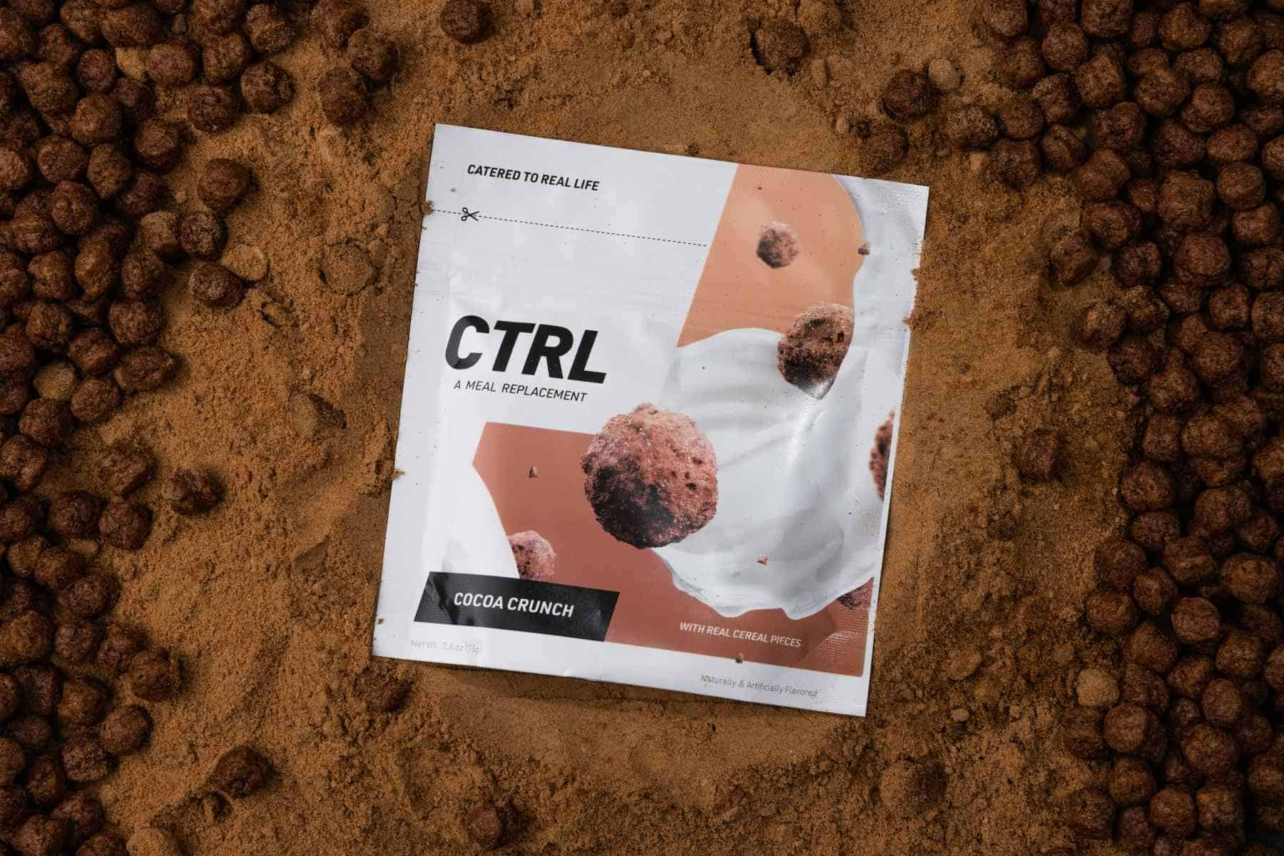 A packet of CTRL Cocoa Crunch meal replacement powder sits on a cocoa-colored surface surrounded by round cereal pieces.