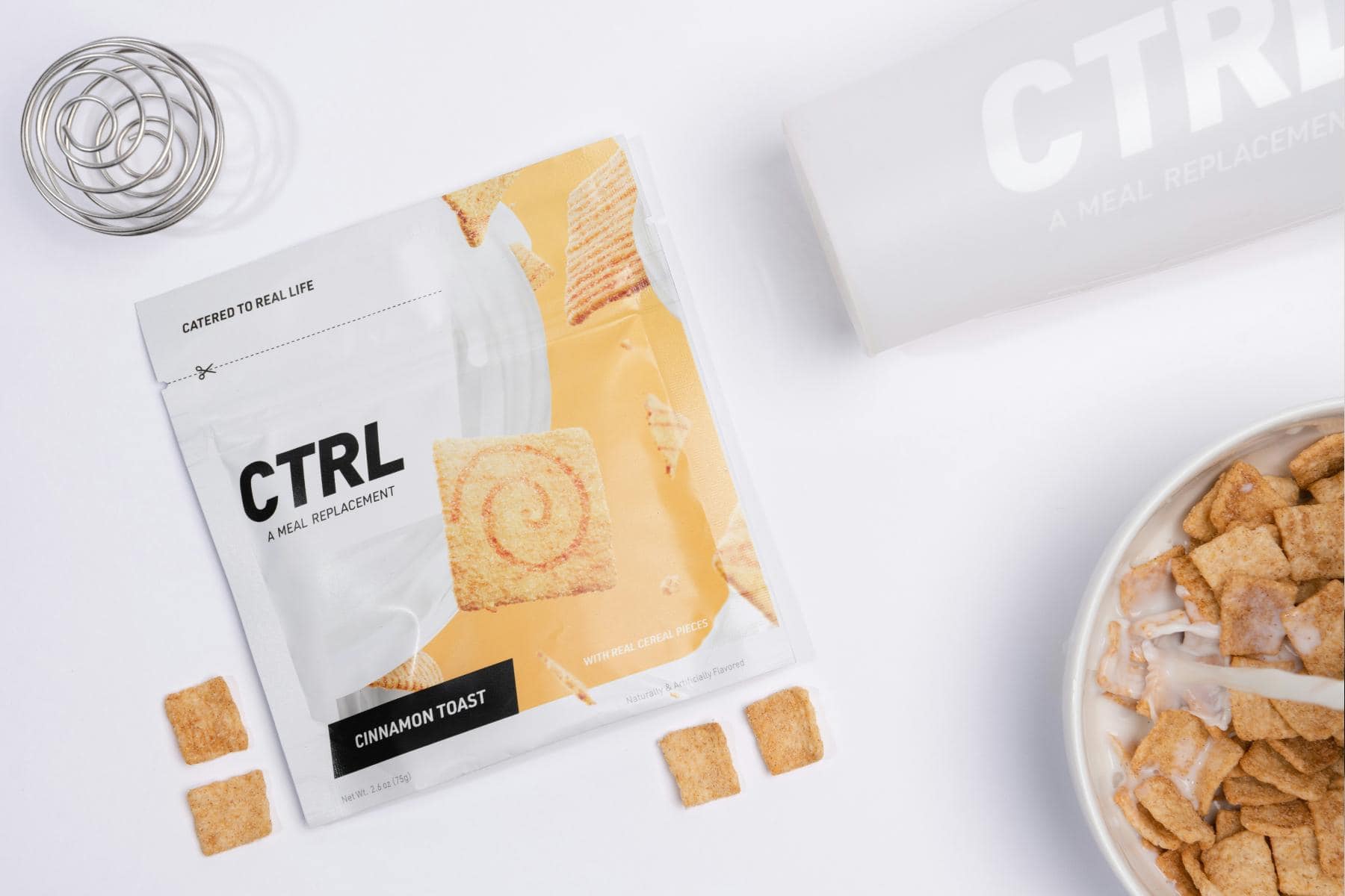 CTRL Cinnamon Toast meal replacement packet, loose cereal squares, a metal shaker ball, a shaker bottle, and a bowl of milk with cereal are arranged on a white surface.