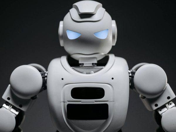 A close-up of a white humanoid robot with blue illuminated eyes, metallic joints, and a screen on its chest, against a dark background.