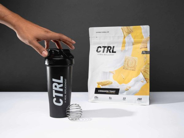 A hand holds a black CTRL shaker bottle next to a pouch of CTRL Cinnamon Toast meal replacement powder with a metal mixing whisk in front.