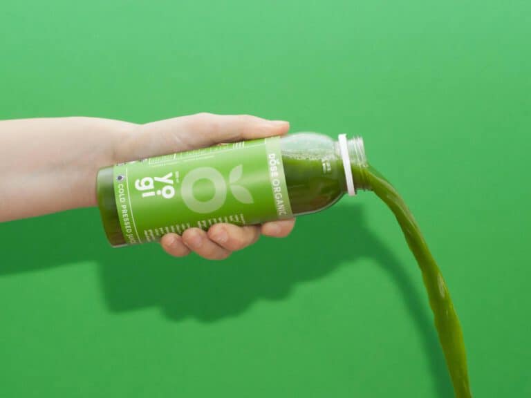 A hand tilts a green bottle of organic juice, pouring the liquid out against a solid green background.