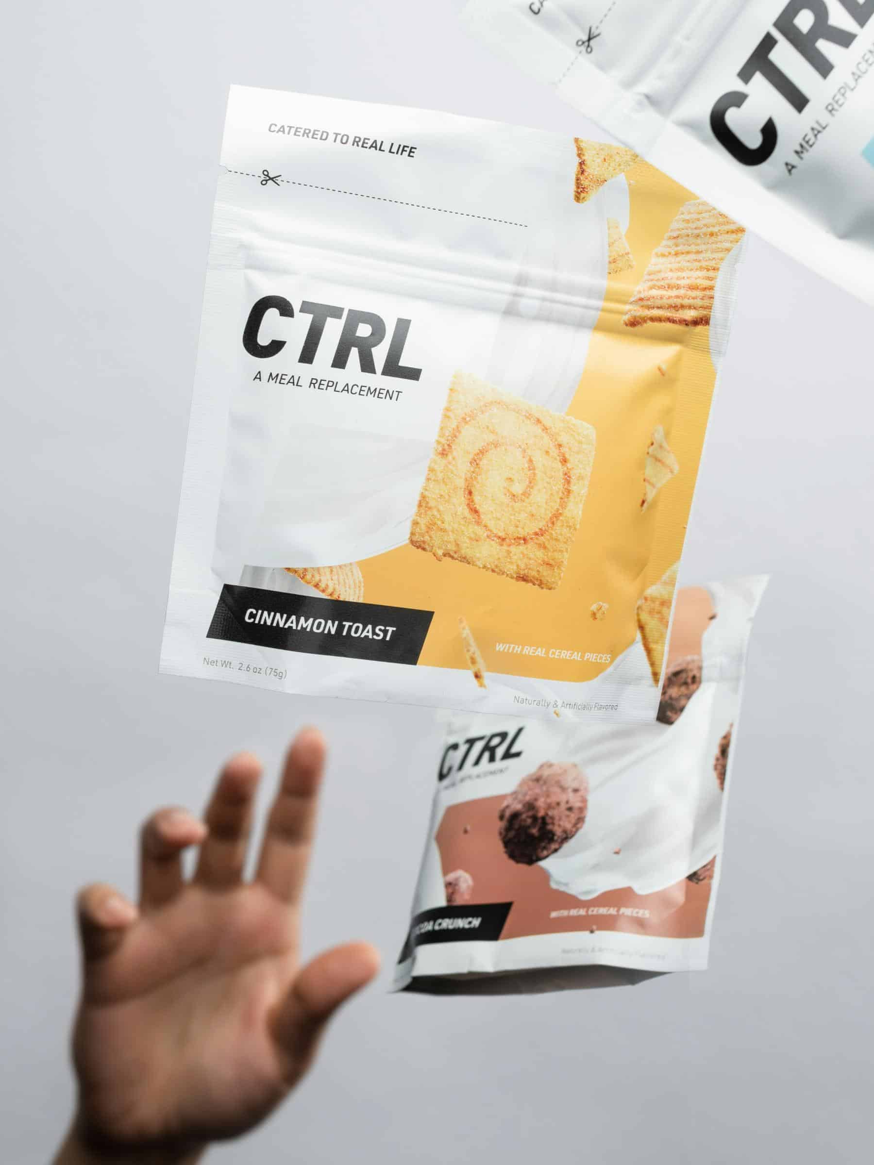 A hand reaches toward two floating CTRL meal replacement packets, one labeled “Cinnamon Toast” and the other “Cocoa Crunch,” against a plain background.