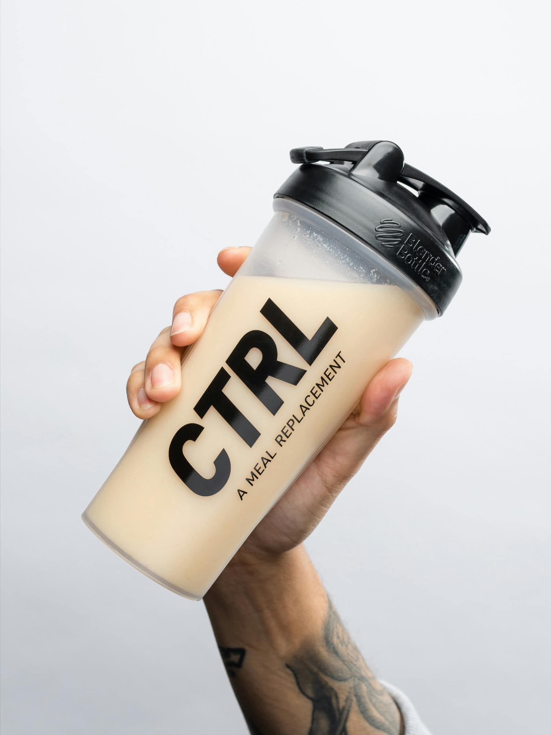 A hand holding a shaker bottle labeled “CTRL A Meal Replacement” filled with a light beige liquid, against a plain light background.