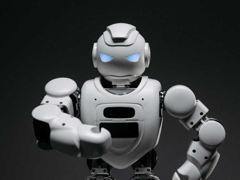 White humanoid robot with glowing blue eyes is shown against a dark background, raising one arm forward in a gesture. The robot has a futuristic and mechanical appearance.