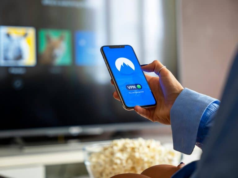 Person holding a smartphone with a VPN app open, seated in front of a TV and a bowl of popcorn.